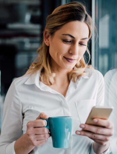 Une femme avec une tasse bleue à la main regarde son smartphone